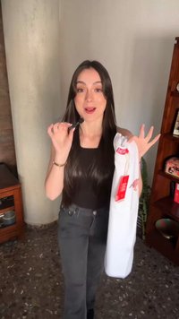 Γυναίκα με μακριά σκούρα μαλλιά κρατάει ένα λευκό αμάνικο τοπ με ετικέτες και ρυθμίζει ένα μικρόφωνο κοντά στο στόμα της μέσα σε εσωτερικό χώρο.