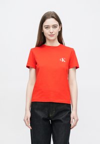 Κόκκινο βαμβακερό t-shirt με κοντά μανίκια, στρογγυλή λαιμόκοψη και λευκό λογότυπο "ck" στο αριστερό μέρος του στήθους. Συνδυασμένο με σκουρόχρωμο τζιν παντελόνι.