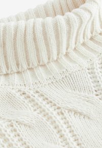 Hellbeiger Strickpullover mit einem gerippten Kragen. Die Textur ist mit Kabelmustern und offenen Maschen gestaltet, was einen schichtenartigen Look verleiht.