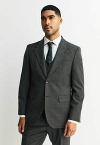 Traje gris a medida con una tela texturizada, cierre de un solo botón y solapas en pico. Combinado con una camisa blanca y una corbata con estampado.