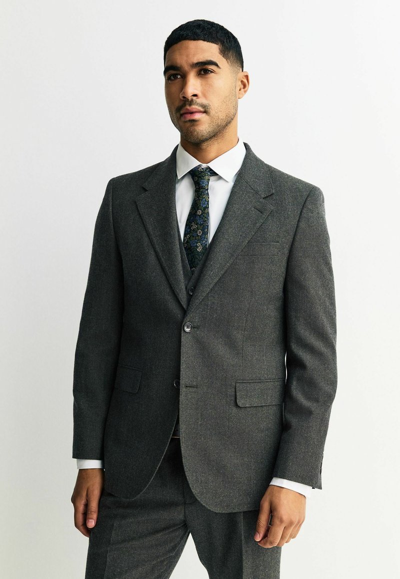 Traje gris a medida con una tela texturizada, cierre de un solo botón y solapas en pico. Combinado con una camisa blanca y una corbata con estampado.