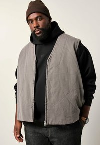 Graue, gestepptes Weste mit glatter Textur, ausgestattet mit einem Reißverschluss und kreisförmigen Steppmustern, lässig über einem schwarzen Hoodie getragen.