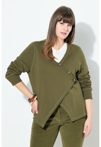 Donna con capelli castani lisci che indossa un cardigan avvolgente verde oliva con bottoni in tartaruga e pantaloni abbinati, mentre posa con una mano sulla vita.