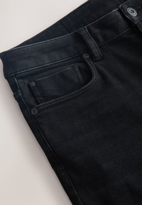 Schwarze Denim-Jeans Nahaufnahme, die Bund, Gürtelschlaufen, Vordertasche und Münztasche mit Nieten-Details auf einer hellen Oberfläche zeigen.