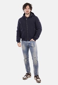 Giacca blu navy con zip e cappuccio, dotata di tasche laterali e polsini a costine, abbinata a jeans chiari strappati e sneakers marroni.