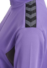 Fioletowa kurtka sportowa z teksturowanej tkaniny, czarnym panelem akcentującym i detalami w wzór chevron na ramionach. Posiada wysoki kołnierz.