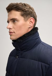 Giacca imbottita blu navy con alto collo e rivestimento in pile nero. Presenta una chiusura con bottoni e una texture trapuntata sul tessuto.