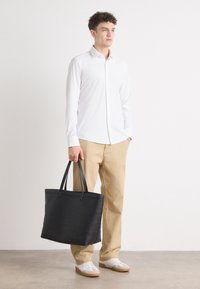 Svart tote bag med to håndtak, glatt tekstur, rektangulær form, sammen med en hvit skjorte og beige bukser, på en person som står mot en lys bakgrunn.