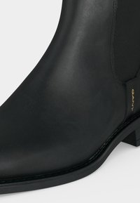 Gros plan d'une bottine en cuir noir avec un panneau latéral élastique et un petit talon bloc, avec le logo doré GANT près de la semelle.