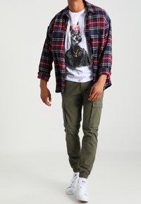 Man med vit grafisk t-shirt som har en hund i jacka, röd och marinblå rutig skjorta, olivgröna cargo-byxor och vita sneakers, står på vit golv.