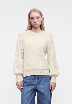 Jeune femme aux cheveux blonds lisses portant un pull en maille torsadée crème et un jean bleu, debout devant un fond blanc uni.