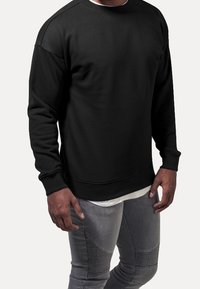 Felpa nera girocollo, realizzata in tessuto liscio, con polsini e orlo elasticizzati, indossata sopra una camicia bianca, abbinata a jeans grigi con texture.