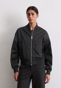 Woman with dark hair in a black zip-up bomber jacket and black pants standing against a plain light wall, facing forward.