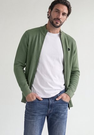 Mann trägt grünen Reißverschluss-Pullover über weißem T-Shirt und blauen Jeans, steht mit den Händen in den Taschen vor hellem, einfarbigem Hintergrund.