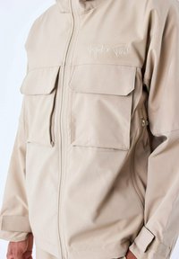 Chaqueta beige con cremallera fabricada en tejido texturizado, que presenta dos grandes bolsillos frontales y detalles de costura. "Project x Paris" está bordado en el pecho.