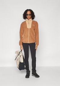 Brun fleecejacka med en beige frontpanel, dragkedjefickor, svarta byxor och svarta vandringsskor. Bär en ljusfärgad ryggsäck.