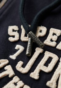 Sort zip-up hoodie med kremfarvede broderede bogstaver og sorte snore med metalanhæng, der er indgraveret med "SUPERDRY."