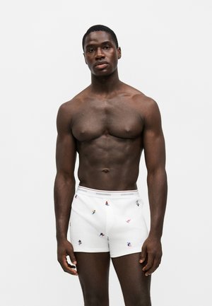 Homme musclé à la peau foncée debout, portant un boxer blanc avec de petits motifs colorés de skieurs sur un fond uni clair.