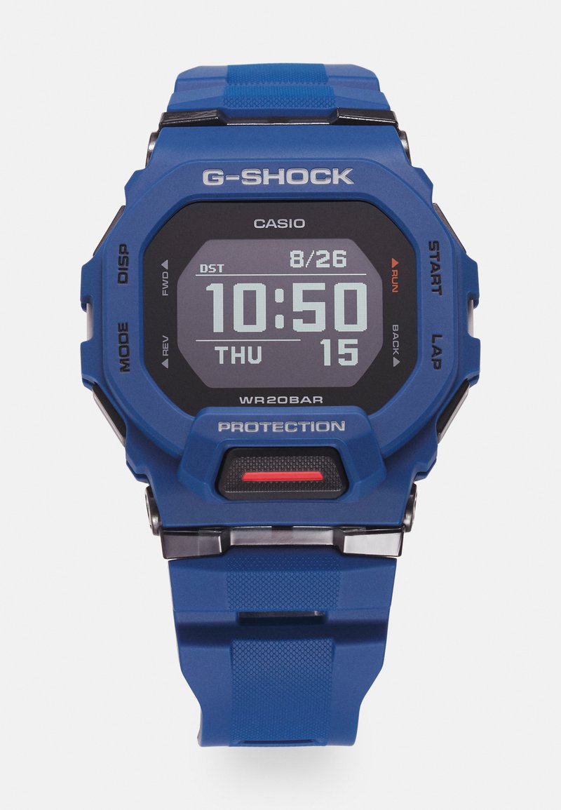 Blå G-Shock-klocka med digital display, funktioner inkluderar tid, datum och stoppur. Texturerad gummirem och röd accentknapp.