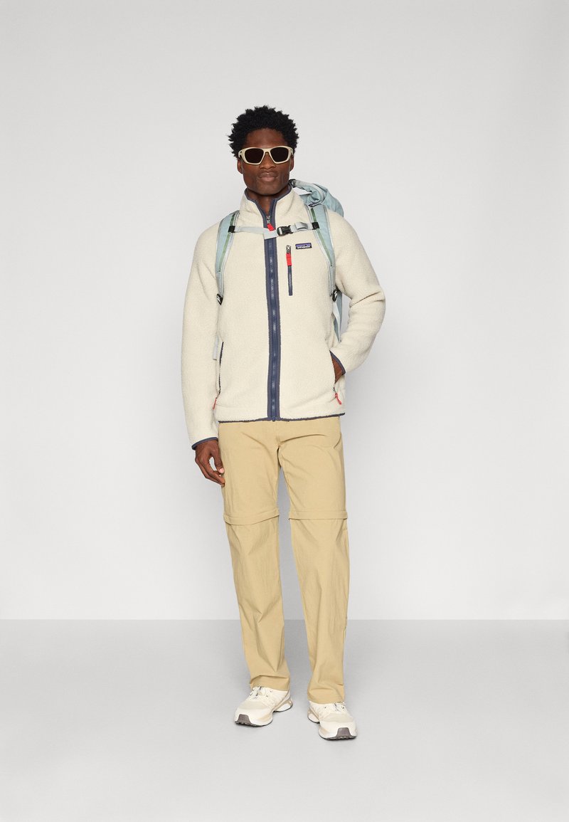 Giacca in fleece color avorio con zip completa e rifiniture blu, abbinata a pantaloni beige convertibili e sneakers bianche. Gli accessori includono uno zaino e occhiali da sole.