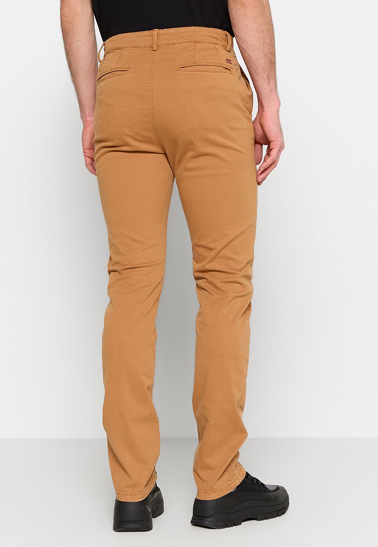 Mężczyzna w wąskich, camelowych spodniach i czarnych sneakersach, ukazany od tyłu na tle białego tła.