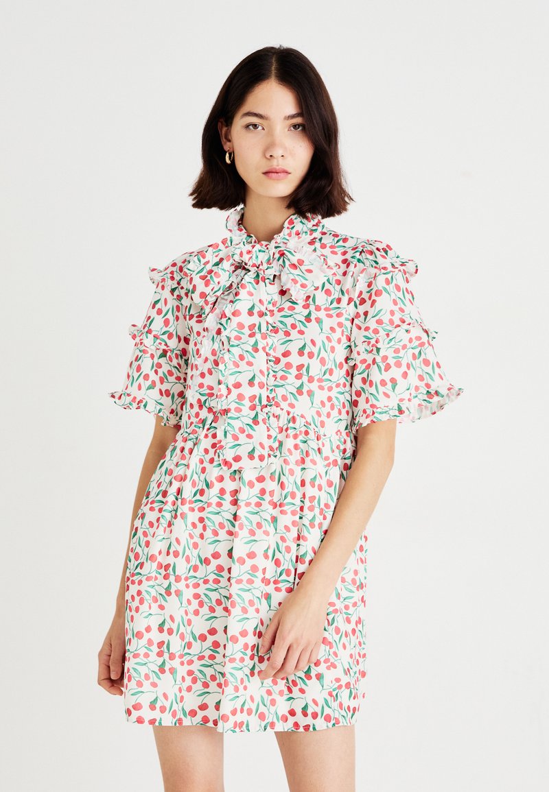 Vestido floral con fondo blanco, que presenta cerezas rosas y hojas verdes, mangas con volantes y detalle de cuello con lazo.