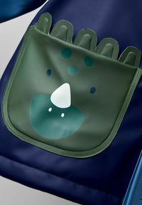 Mochila de tela azul marino con un bolsillo verde que presenta la cara de un animal de dibujos animados, incluyendo un hocico redondeado, ojos y patrones de puntos.