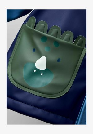 Tumedast kangast seljakott, millel on roheline tasku koos karikatuurilaadse looma näo kujutisega, sealhulgas ümar kuju nina, silmad ja punktmustrid.