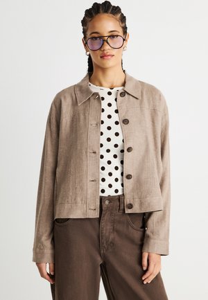 Woman wearing light brown buttoned jacket over white polka-dot shirt with brown pants and gradient-tinted sunglasses, against plain background.