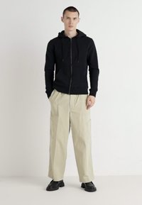 Black zip-up hoodie with hood, paired with light beige cargo pants featuring side pockets, and black slip-on shoes. Smooth texture overall.