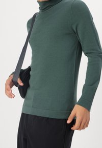 Pull vert à manches longues avec col roulé, confectionné dans un matériau doux, doté d'une coupe ajustée et d'un col côtelé, accompagné d'un sac bandoulière noir.