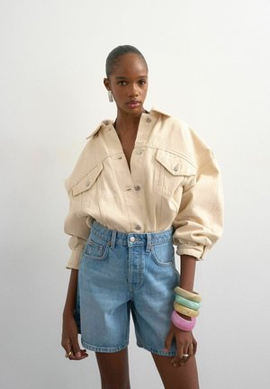 Femme portant une veste beige oversize à boutons, un short en denim bleu taille haute, des bracelets colorés et épais, et des boucles d'oreilles rectangulaires sur fond blanc.