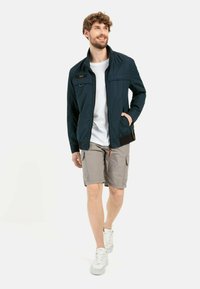 Marineblau leichte Jacke mit einem Reißverschluss vorne, zwei seitlichen Taschen und einem gerippten Saum, kombiniert mit beigen Cargo-Shorts und weißen Sneakern.