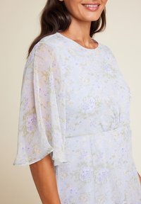 Blomsterkjole med gjennomsiktige, klokkeformede ermer i lys blått med lilla og grønne detaljer, delikate blomstermønstre, og en innsnevret midje.