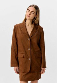 Blazer en daim marron avec col à revers, fermeture à bouton simple et deux poches avant. Texture lisse, design ajusté.