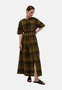 Robe maxi à manches courtes, motif à carreaux en olive et noir, taille ceinturée, fermeture à boutons sur le devant, fente sur le côté, associée à des sandales noires.
