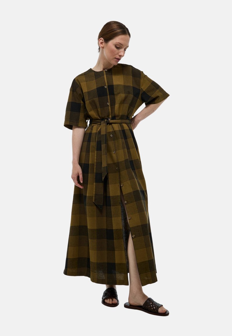 Robe maxi à manches courtes, motif à carreaux en olive et noir, taille ceinturée, fermeture à boutons sur le devant, fente sur le côté, associée à des sandales noires.