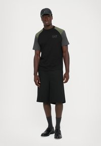 Man wearing a black and olive raglan sleeve T-shirt, black long shorts, gray socks, black loafers, and a black leather cap, standing against white background.
