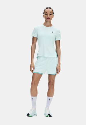 Athletische Person steht, trägt ein hellmintfarbenes kurzärmeliges Shirt, passende Shorts, weiße Socken und weiße Laufschuhe mit grünen Akzenten.