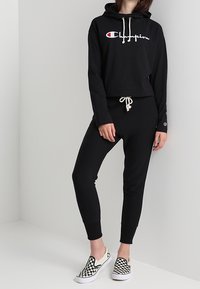 Zwarte cropped hoodie met logo aan de voorkant, gecombineerd met zwarte aansluitende sweatpants. Gecombineerd met geruite slip-on schoenen.
