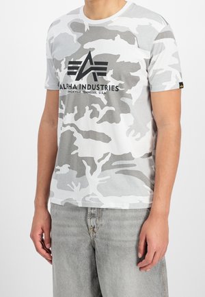 Mand iført en grå og hvid camouflage Alpha Industries T-shirt og lysegrå denimjeans, stående med armene afslappede.