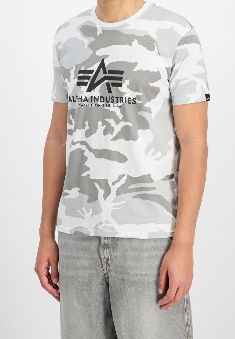 Homme portant un t-shirt Alpha Industries camouflage gris et blanc et un jean en denim gris clair, debout les bras détendus.