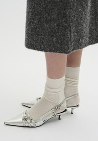 Scarpe décolleté metalliche argento con punta affilata, caratterizzate da accenti a strappo e tacco piccolo, abbinate a calze a coste beige chiaro.
