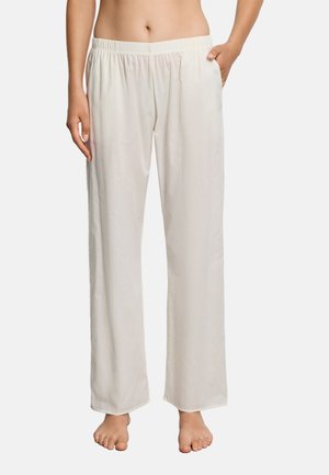 Frau trägt lockere, hoch taillierte cremefarbene Hose mit elastischem Bund, steht barfuß vor einfachem Hintergrund.