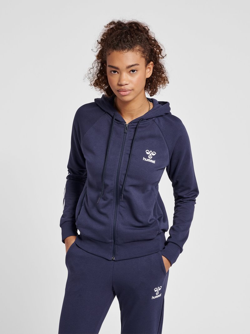 Młoda kobieta z kręconymi włosami ubrana w granatową bluzę z zamkiem marki hummel i pasujące spodnie dresowe, z rękami w kieszeniach.