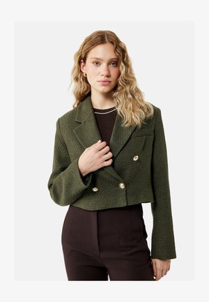 Frau mit welligem blondem Haar, die einen olivgrünen, kurz geschnittenen, zweireihigen Blazer mit goldenen Knöpfen und dunkle hoch taillierte Hosen trägt.