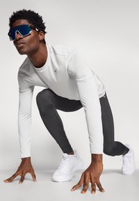 Sportists baltā garajocā, melnās leggingās un baltās skriešanas kurpēs. Valkājot atstarotā zila sporta brilles, noliektā pozīcijā.