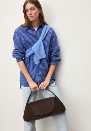 Camisa de rayas azules y blancas, suéter azul atado al cuello, jeans azul claro, sosteniendo un bolso marrón de ante con forma curva y cierre de solapa.