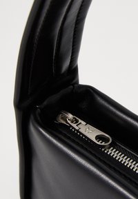 Črna usnjena torba z gladko teksturo, ki ima zgornjo ročico in srebrno zadrgo s vidnimi zobmi in podrobnostmi logotipa.
