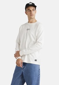 Long-sleeve white T-shirt with ribbed cuffs, featuring a black logo on the chest and a small brand tag on the bottom left. Worn with blue jeans.